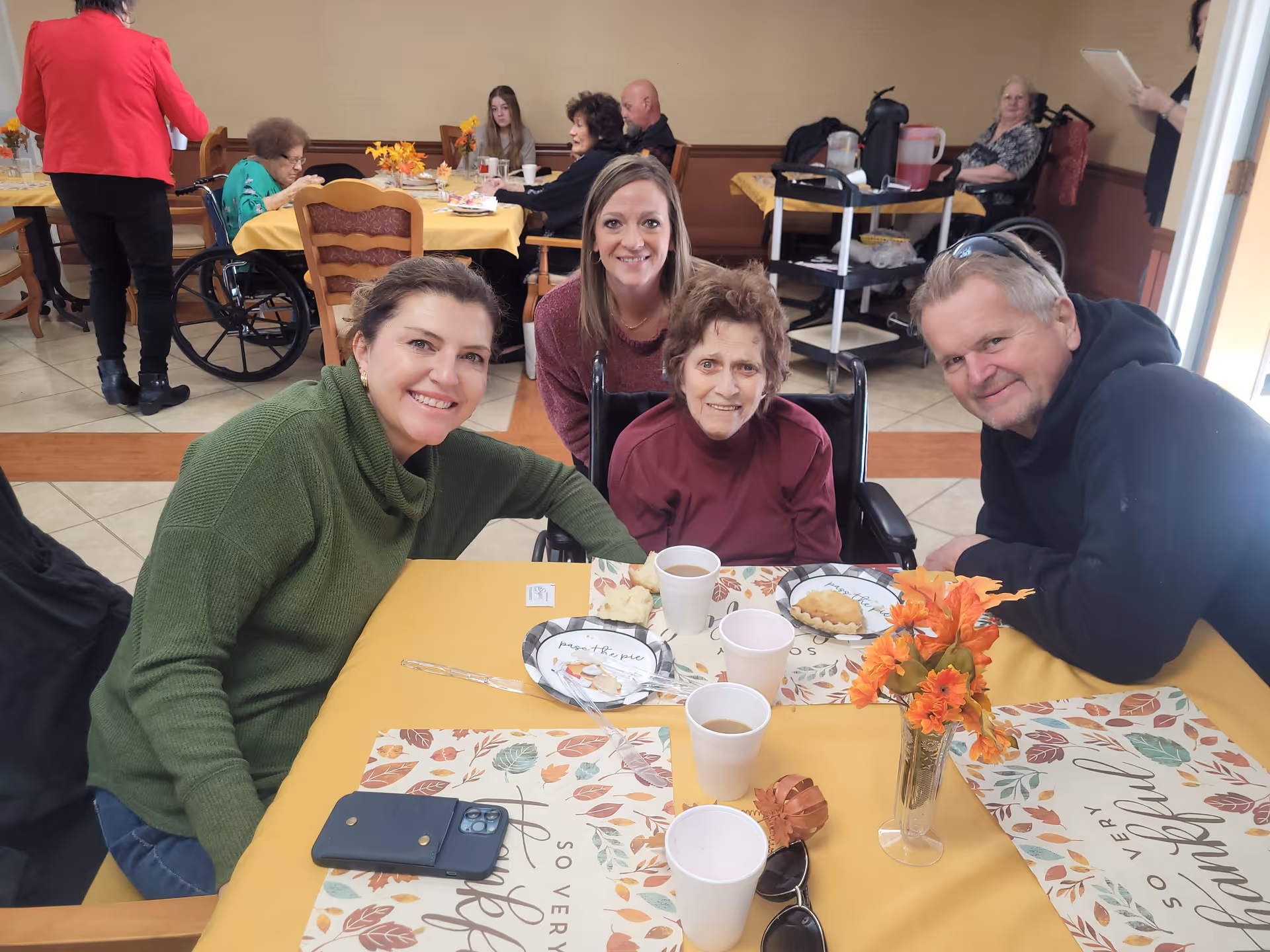 A group of four people, including an elderly woman in a wheelchair, sitting around a table with yellow tablecloths and autumn-themed placemats. They are smiling at the camera in a dining area with other people seated at tables in the background.
