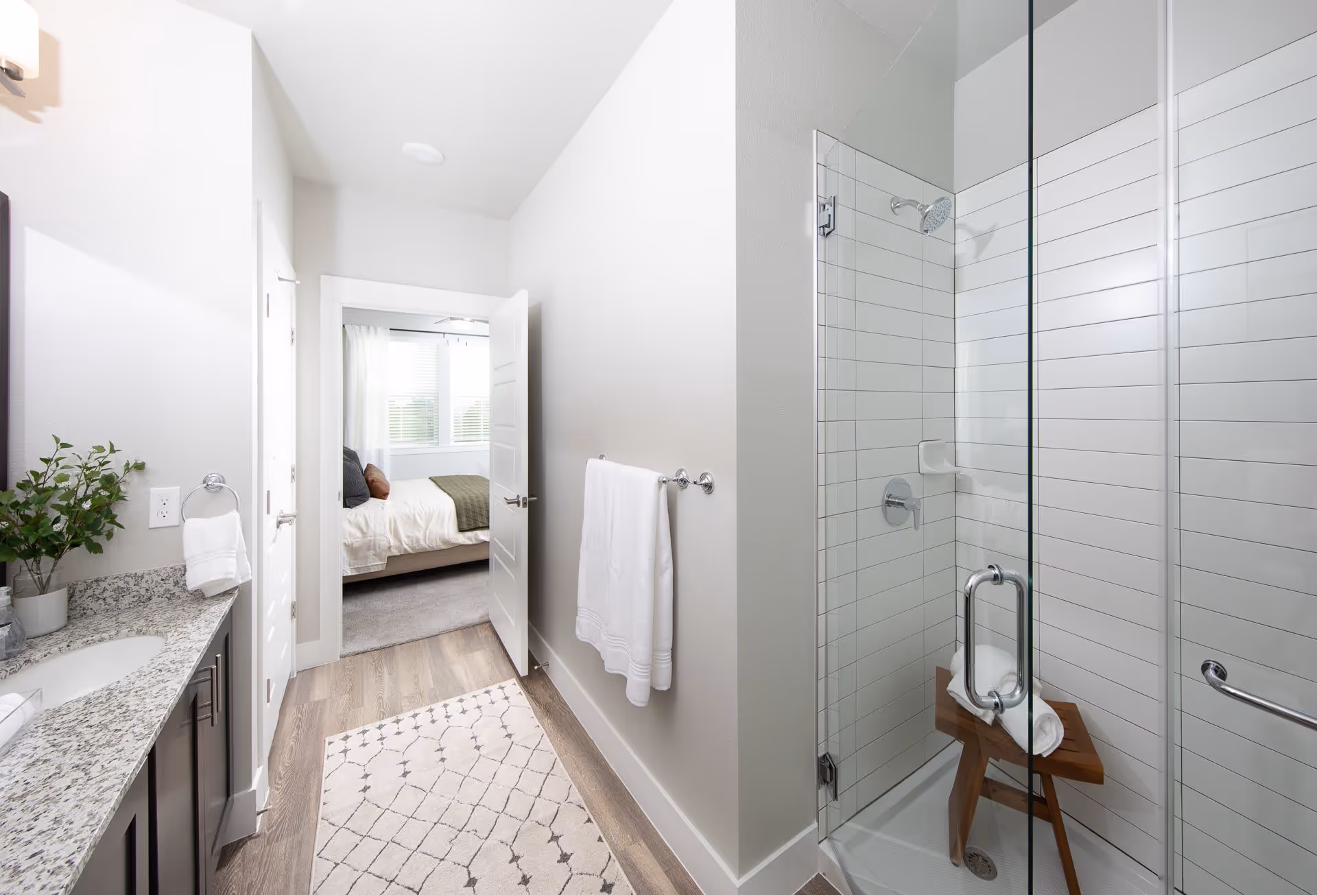 A modern bathroom with a granite countertop and sink on the left, a glass-enclosed shower with white tiled walls and a wooden stool inside on the right, and an open door leading to a bedroom with a bed and windows in the background.