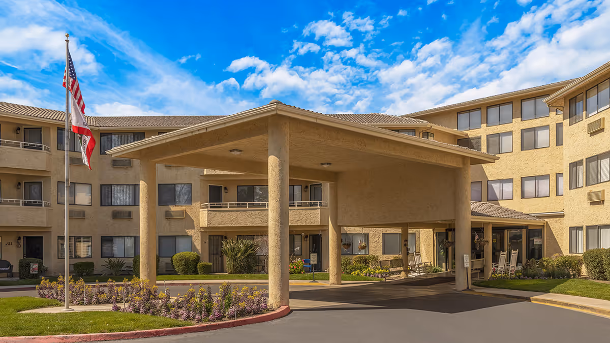 Covered entrance and porte-cochere of a beige multi-story senior living building with flags and landscaped flowerbeds.