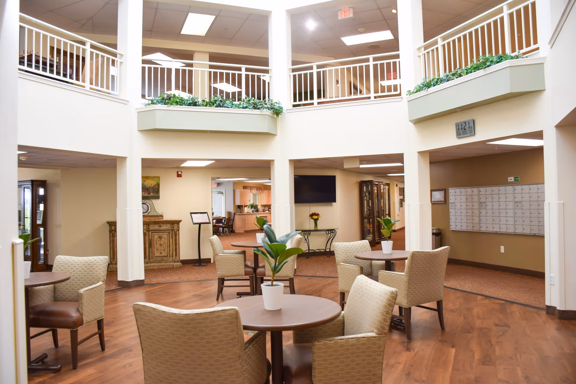 Interior view of a senior living facility common area with round tables and chairs arranged on a wooden floor. There are potted plants on the tables, a TV mounted on the wall, and a set of mailboxes on the right side. The area has a two-story open ceiling with white railings on the upper level and some greenery decoration along the balcony edges.