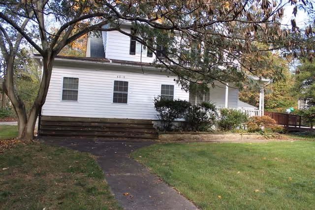 A white two-story house with several windows and a porch, surrounded by green grass, bushes, and trees with autumn leaves. A paved walkway leads up to the house.