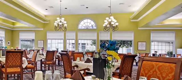 A bright and spacious dining room with multiple tables covered in white tablecloths, set with glasses and napkins. The room features large windows with white blinds, yellow-green walls, ornate chandeliers, and comfortable upholstered chairs with a patterned design. A vase with colorful flowers is visible on one of the tables.