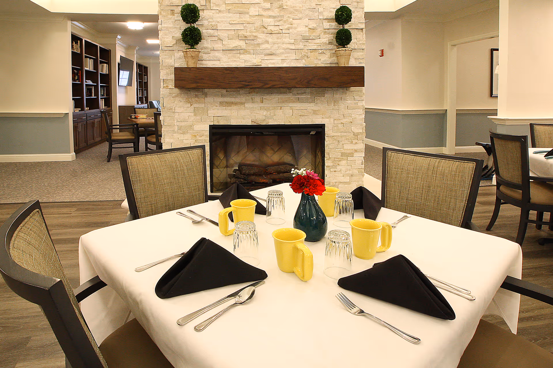 A dining table set for four with yellow mugs, upside-down glasses, black folded napkins, and silverware. A small vase with red and white flowers is in the center. The table is in front of a stone fireplace with a wooden mantel decorated with two small potted plants. The room has beige walls with blue-gray wainscoting and additional seating and bookshelves visible in the background.
