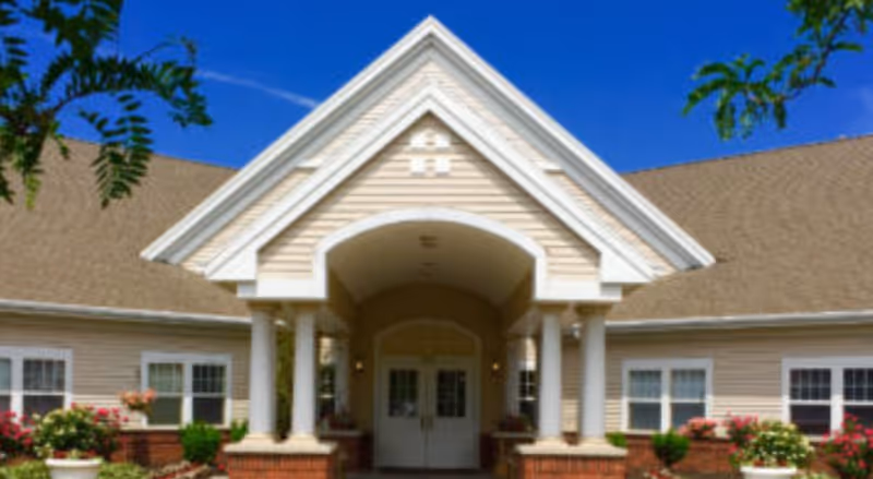 Front entrance of a single-story senior living building with a gabled covered portico, columns, double doors, and landscaping.