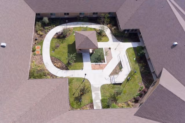 Aerial view of a courtyard within a building complex featuring a central gazebo, curved and straight concrete walkways, green grass, small trees, and landscaped garden beds surrounded by the building's roof.
