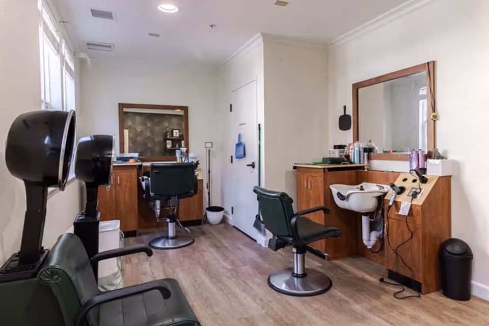 Interior view of a hair salon area within a senior living facility, featuring two green salon chairs, two black hair dryers, a wooden cabinet with a sink, hair care products, and a large mirror on the wall.