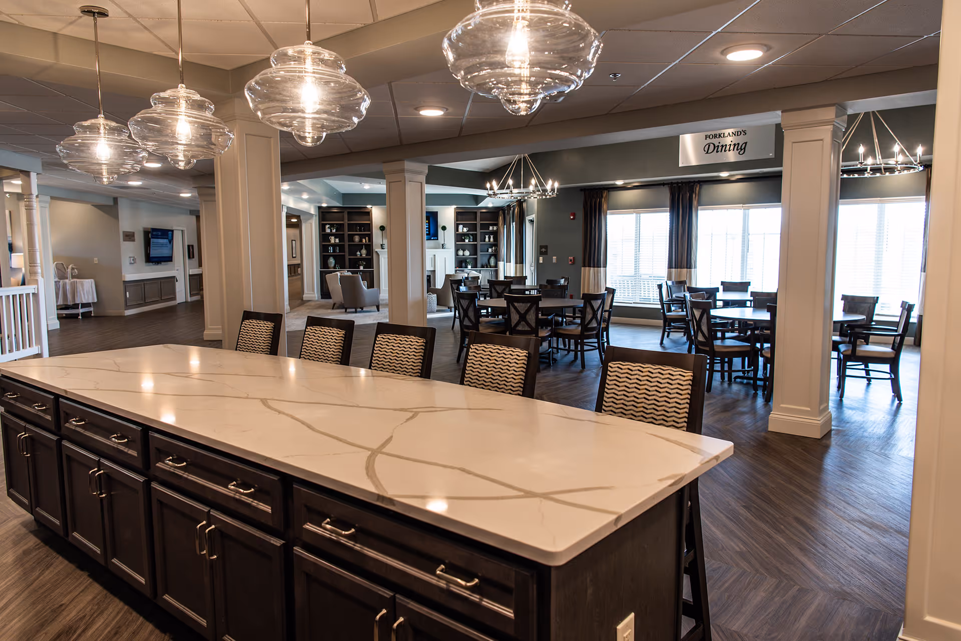 Interior view of a senior living facility dining area with a large marble countertop island in the foreground surrounded by chairs. The background shows multiple dining tables and chairs, large windows with curtains, and a sign that reads 'Forkland's Dining'. The space is well-lit with modern hanging light fixtures and has a warm, inviting atmosphere.