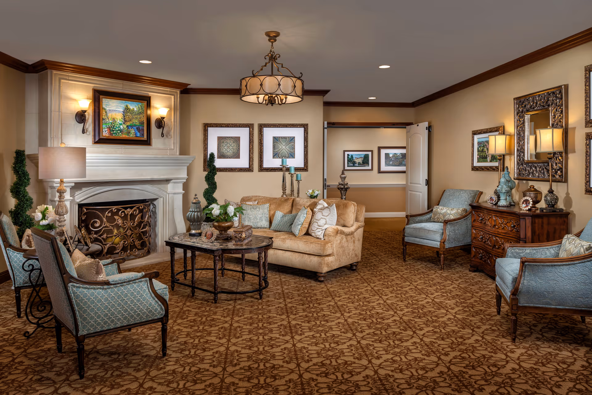 A cozy living room area in The Kensington Sierra Madre featuring a beige sofa with multiple cushions, two blue upholstered armchairs, and two additional chairs with patterned upholstery. The room has a decorative fireplace with a painting above it, a round coffee table with decorative items, a wooden chest of drawers with lamps and ornaments, and framed artwork on the walls. The carpet has an intricate pattern, and the ceiling has recessed lighting and a central chandelier.