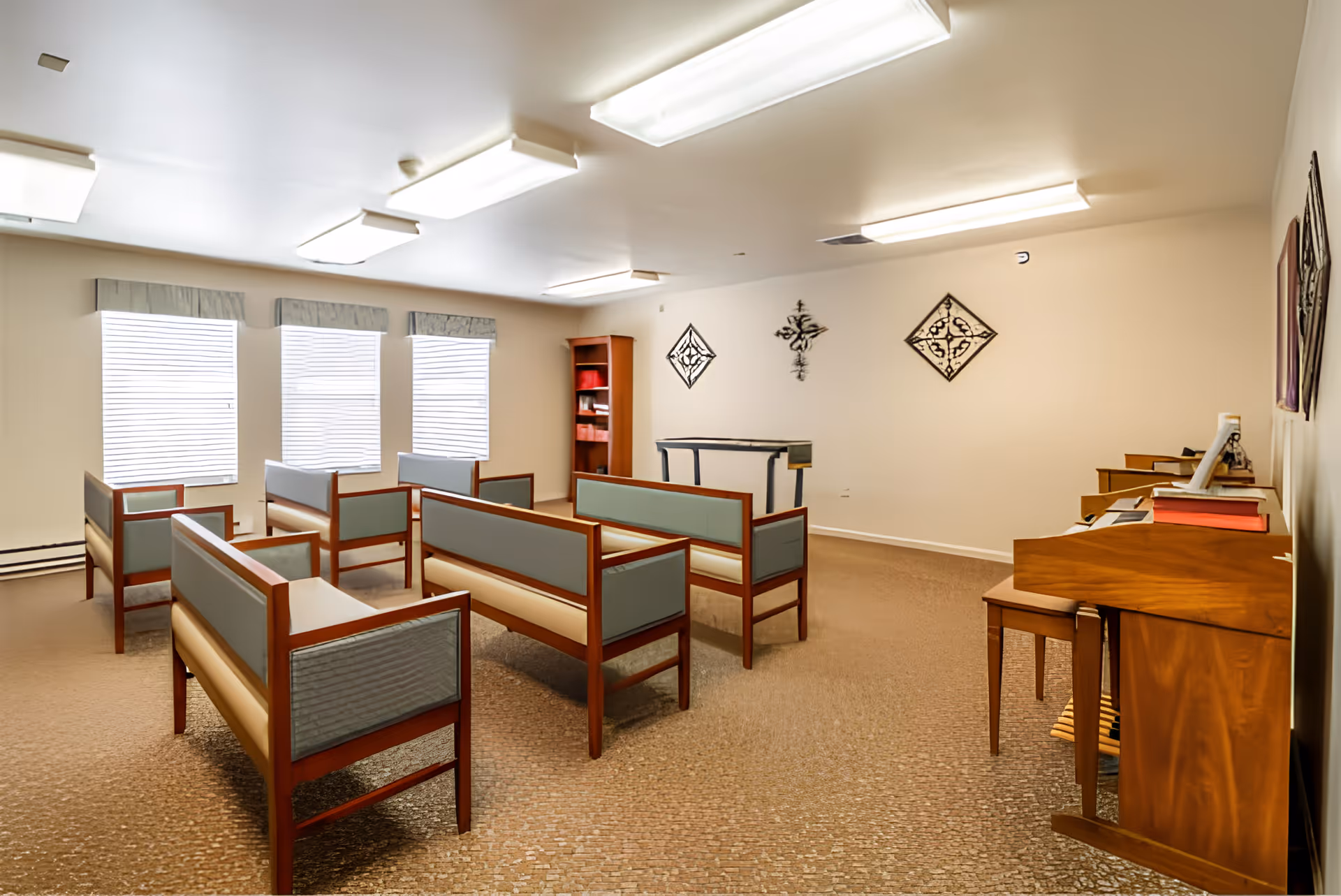 A well-lit room with several wooden benches with light blue cushions arranged in rows facing a small podium. The room has three large windows with blinds and valances, a bookshelf in the corner, and decorative wall hangings. There is also a wooden piano with a bench on the right side of the room.