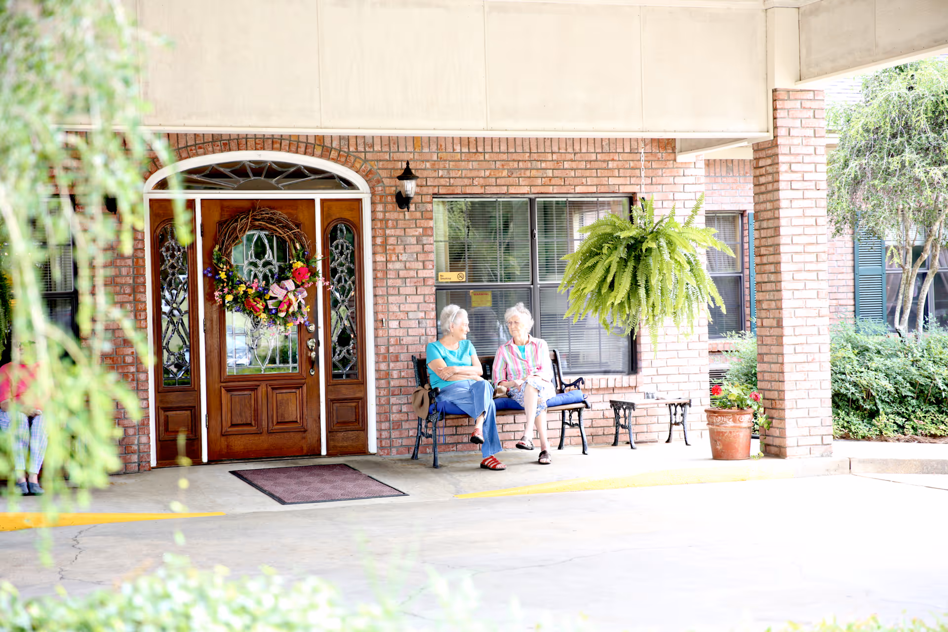 Two elderly women sit on a bench outside the brick front entrance of a senior living facility with double wooden doors, a wreath, and a hanging fern.