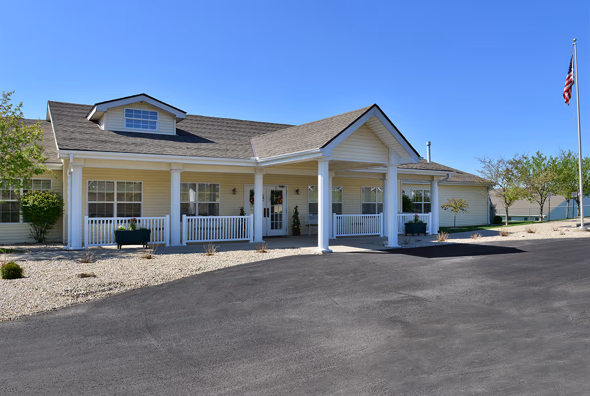 Single-story senior living facility front with a covered entrance, white columns, small porch and an American flag under a clear blue sky.