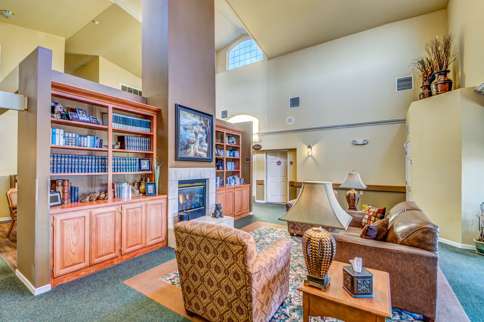 Spacious senior living common room with built-in bookshelves, a fireplace, high ceilings, and a seating area with chairs and lamps.