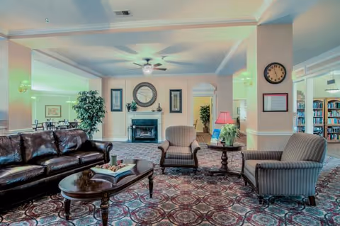 A spacious living room area with a dark leather sofa, two striped armchairs, and a wooden coffee table with a decorative book and candles. The room features a patterned carpet, a fireplace with a large round mirror above it, framed artwork on the walls, a ceiling fan with lights, and a clock on a pillar. There are plants and bookshelves visible in adjoining rooms.