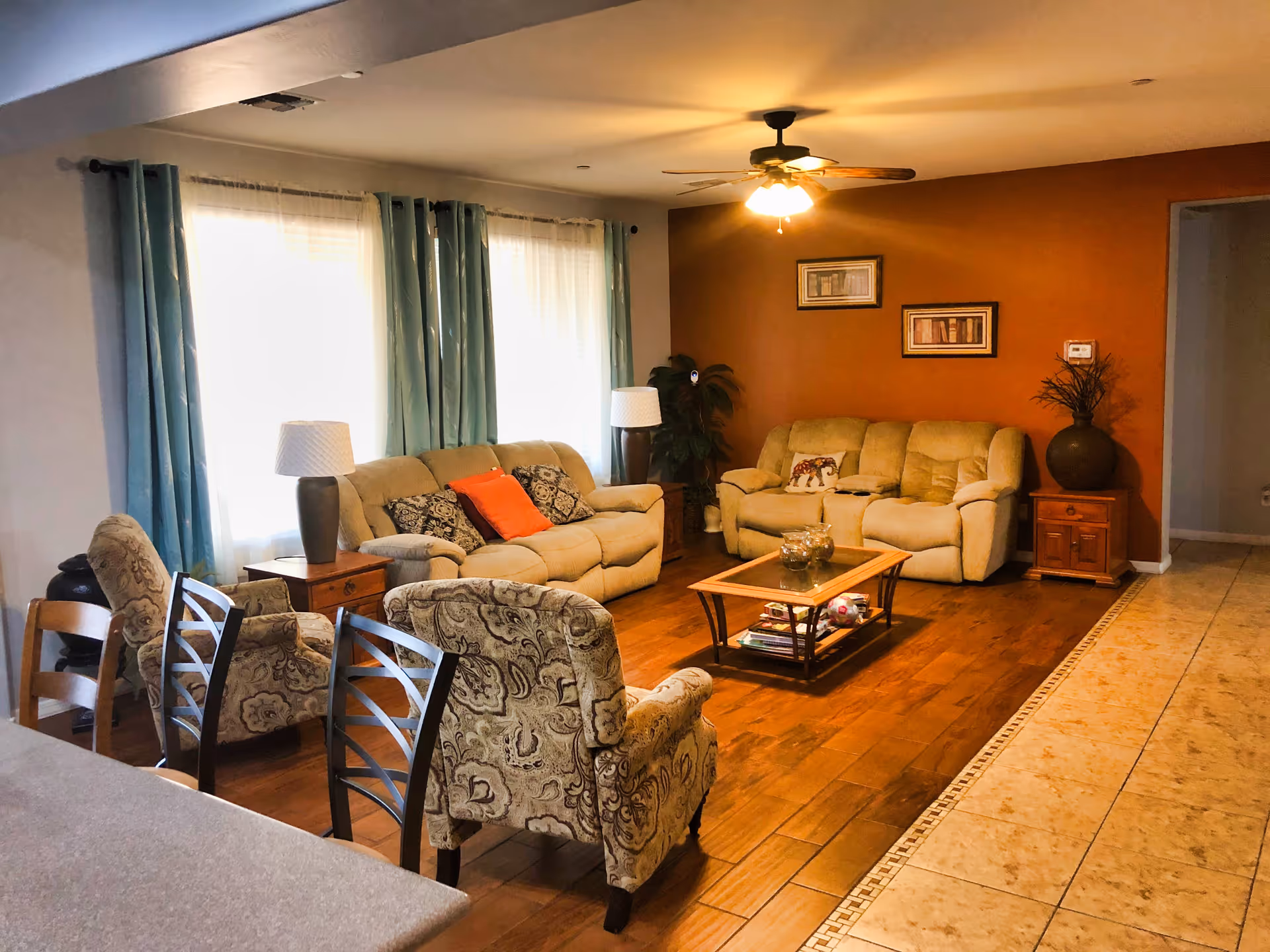A cozy living room with two beige sofas, two patterned armchairs, a wooden coffee table with decorative items, two side tables with lamps, teal curtains covering two windows, a ceiling fan with lights, and framed artwork on a burnt orange accent wall.