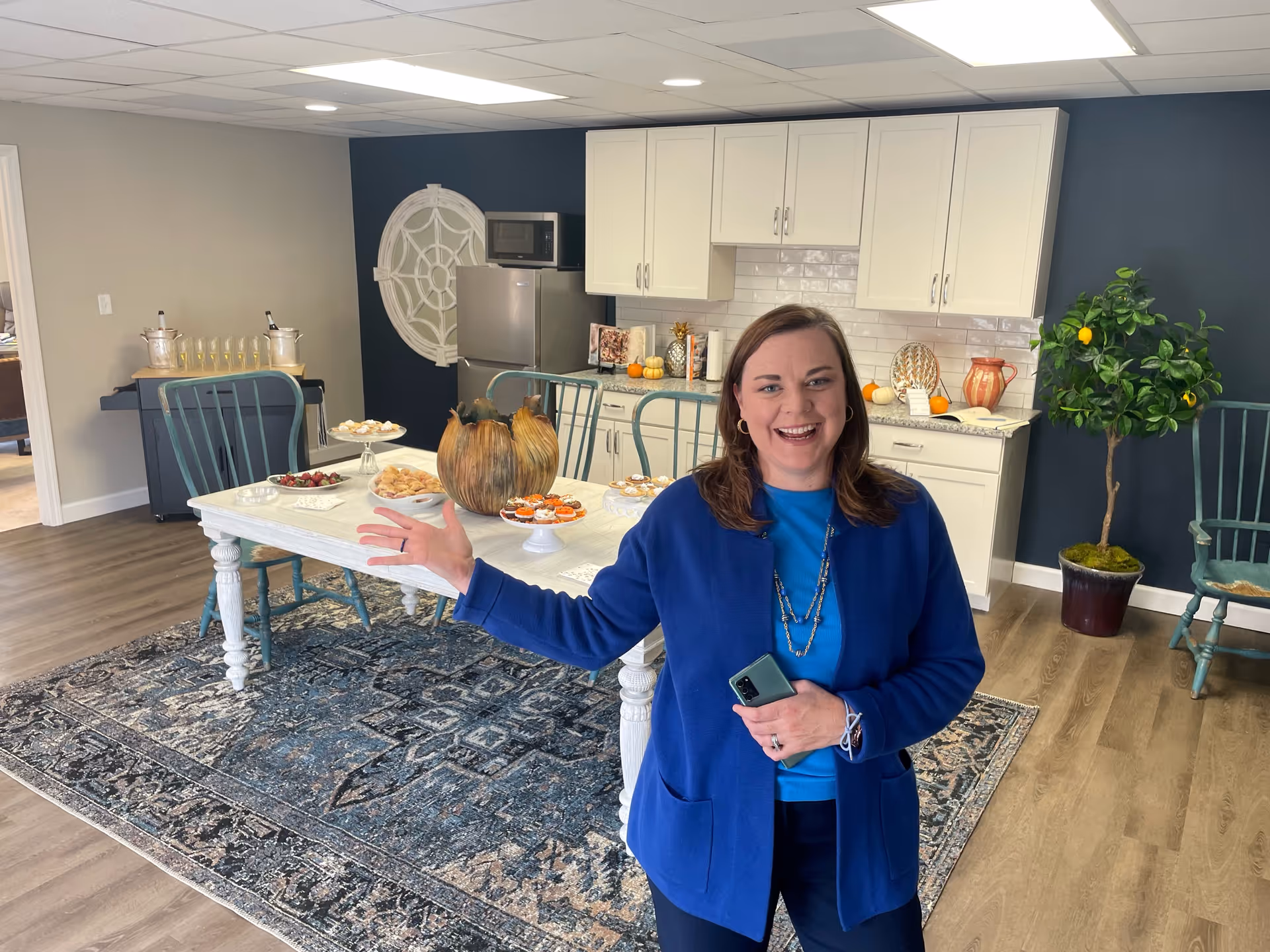 A smiling woman in a blue cardigan stands in a room with a white table set with various snacks and decorative pumpkins. Behind her is a kitchenette with white cabinets, a refrigerator, and a microwave. The room has wooden flooring, a patterned rug, and teal chairs. There is a potted plant with yellow fruit on the right side and a dark accent wall with a decorative white circular wall piece.