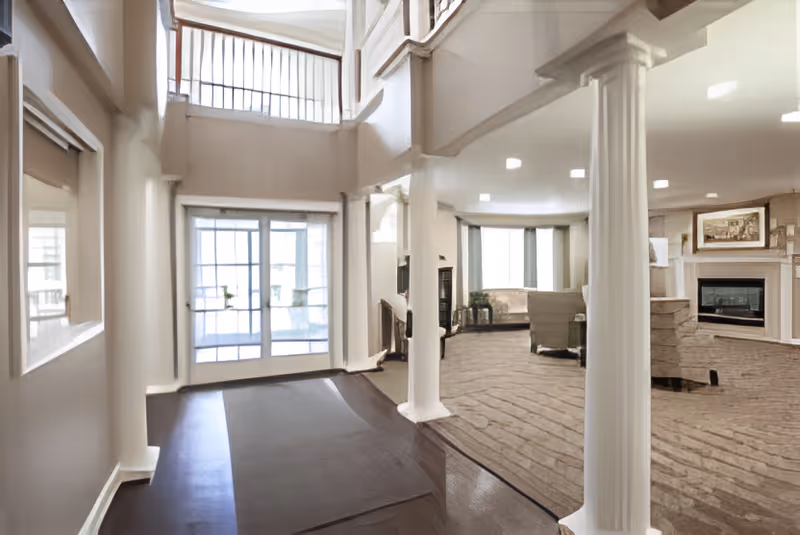 Interior view of a senior living facility lobby area with large windows and glass doors letting in natural light. The space features white columns, a seating area with chairs and a fireplace, and a carpeted floor. The walls and ceiling are painted in neutral tones, creating a bright and welcoming atmosphere.