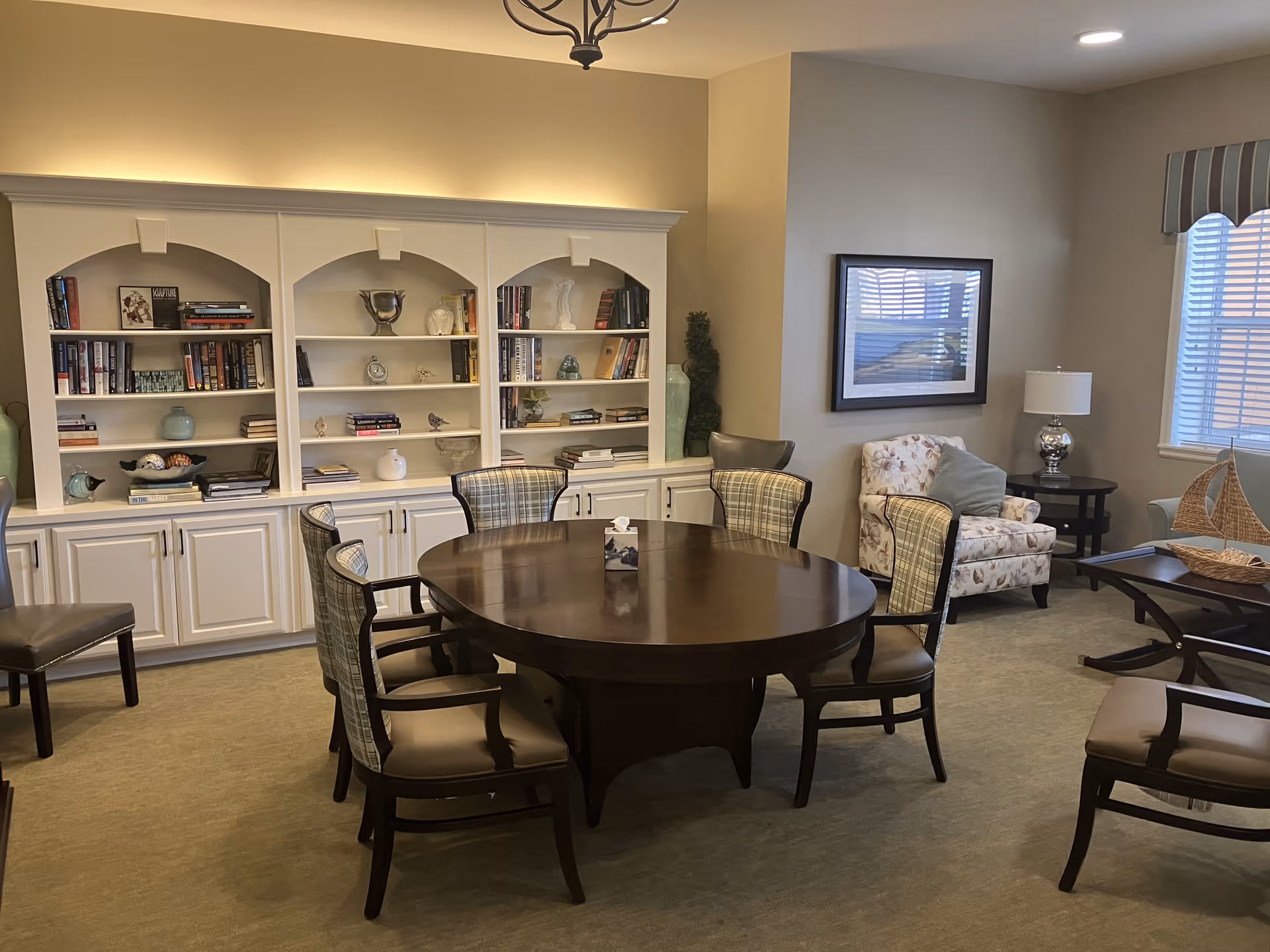 A cozy living room area with a round wooden table surrounded by six cushioned chairs. Behind the table is a white built-in bookshelf filled with books and decorative items. To the right, there is a floral armchair with a green pillow, a side table with a lamp, and a window with blinds and a striped valance. A framed picture hangs on the wall above the armchair.
