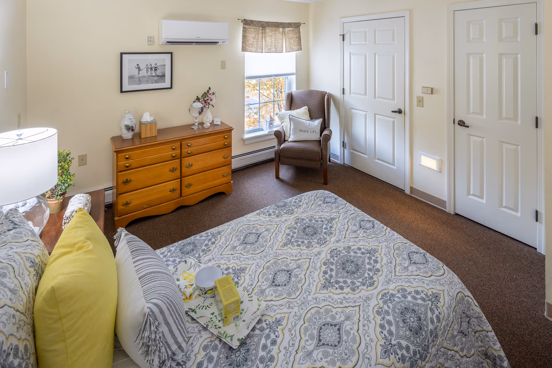 Sunny furnished bedroom with a patterned bed, wooden dresser, armchair by the window, and two white doors.