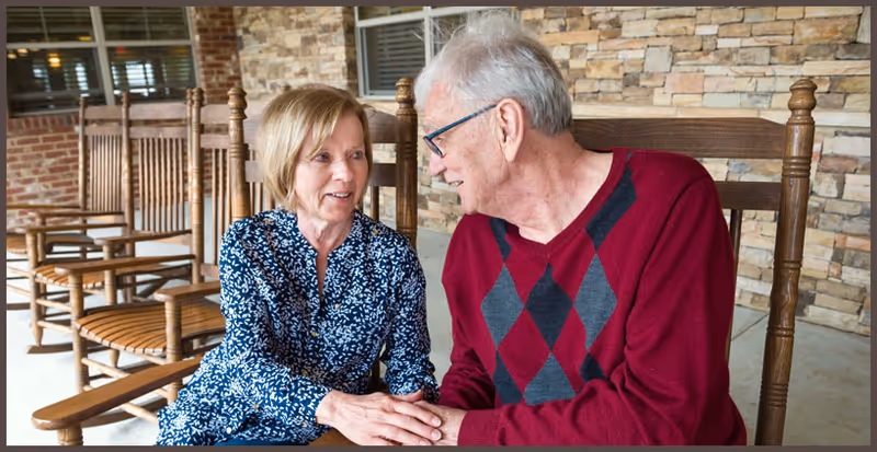 An elderly man and woman sitting on wooden rocking chairs on a porch with a stone wall background. They are holding hands and looking at each other, smiling.