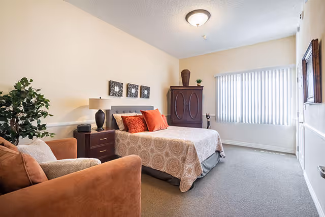 A cozy bedroom with a bed covered in a patterned bedspread and decorated with orange and beige pillows. Next to the bed is a wooden nightstand with a lamp and a small clock. There is a brown armchair with a cushion in the foreground and a tall wooden cabinet in the corner near a window with vertical blinds. The walls are painted light beige and the room is carpeted.