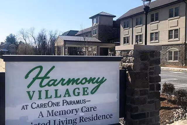 Exterior view of Harmony Village at CareOne Paramus, a memory care assisted living residence, showing the building with multiple windows and a clock tower, along with a large sign in the foreground displaying the facility's name and description.
