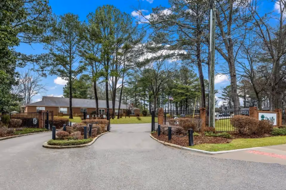 Entrance driveway to Pine Valley Retirement Community with a gated fence, brick pillars, manicured bushes, and tall trees under a blue sky with some clouds.