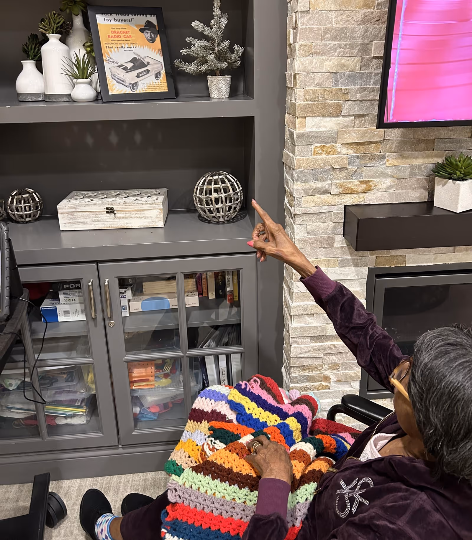 A person seated with a colorful crocheted blanket points toward a gray built-in shelving unit and wall-mounted TV in a living room-style common area.