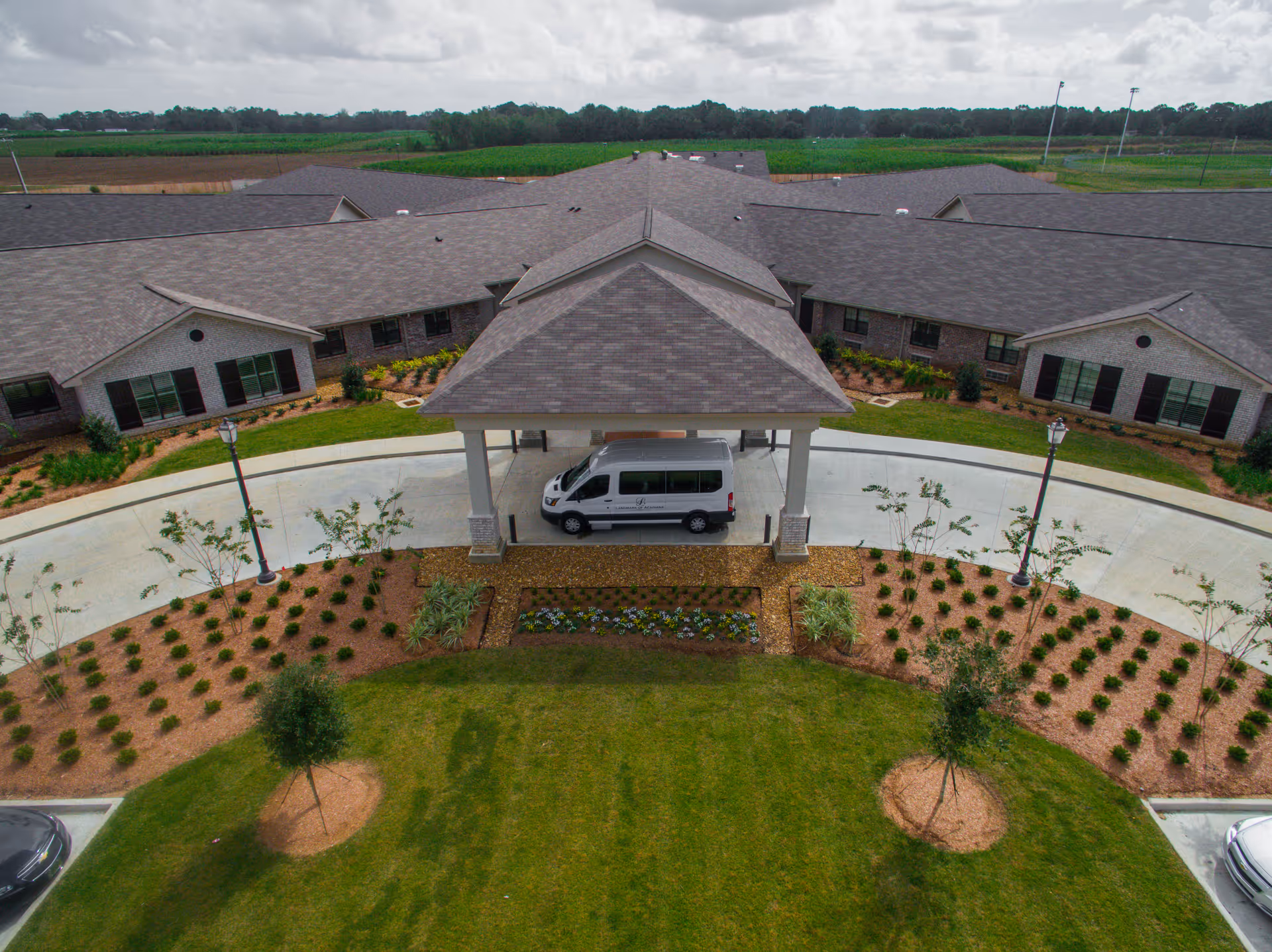 Aerial view of the front entrance of a senior living facility named Landmark Of Acadiana, featuring a covered drop-off area with a white van parked underneath. The building has a gray shingled roof and brick exterior walls, surrounded by landscaped gardens with small trees, shrubs, and flower beds. A circular driveway and street lamps are also visible.