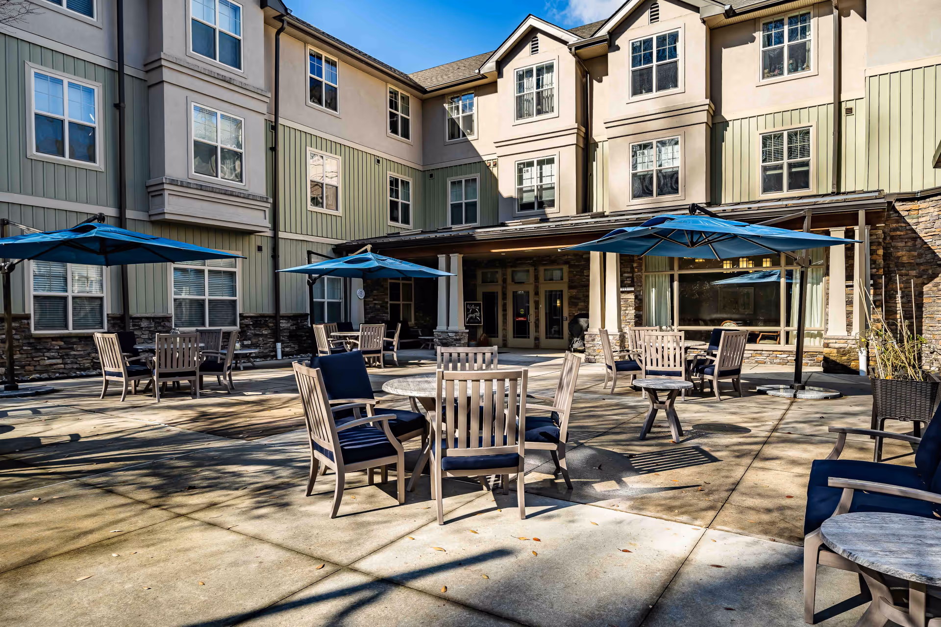 Outdoor patio area of a senior living facility with multiple wooden tables and chairs, each shaded by blue umbrellas. The patio is surrounded by a multi-story building with beige and green siding and numerous windows. The sky is clear and blue.