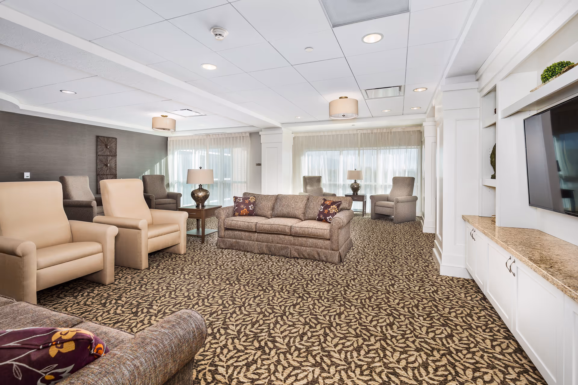 A spacious and well-lit living room area with patterned carpet, beige and gray armchairs, a gray sofa with decorative pillows, side tables with lamps, large windows with sheer curtains, and a wall-mounted flat-screen TV above a built-in cabinet with a granite countertop.