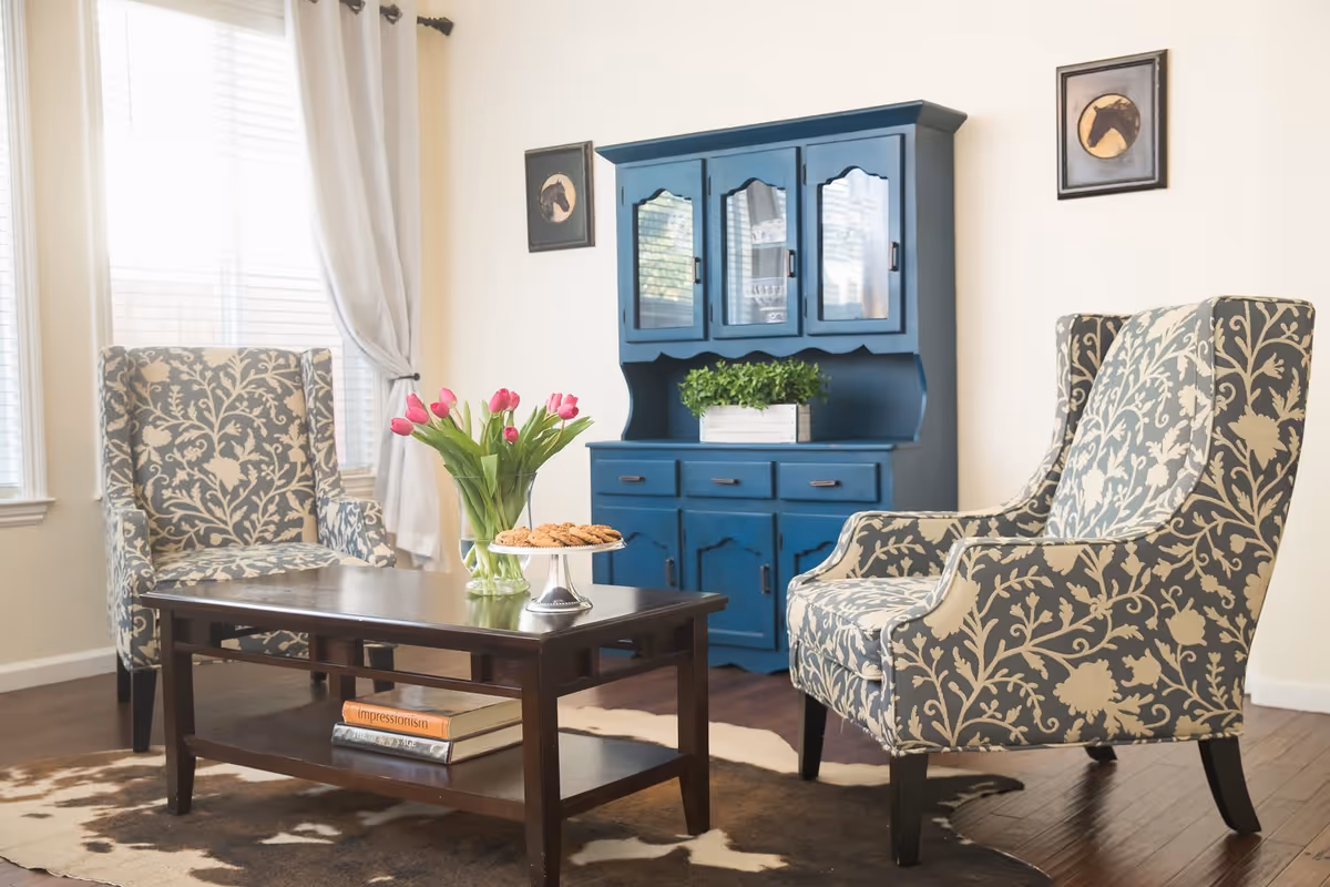 A cozy living room area with two patterned armchairs facing a dark wooden coffee table. On the table, there is a vase with pink tulips and a plate of cookies. Behind the chairs is a blue cabinet with glass doors and a potted plant on top. The room has light-colored walls, two framed pictures of horses, and large windows with white blinds and curtains.