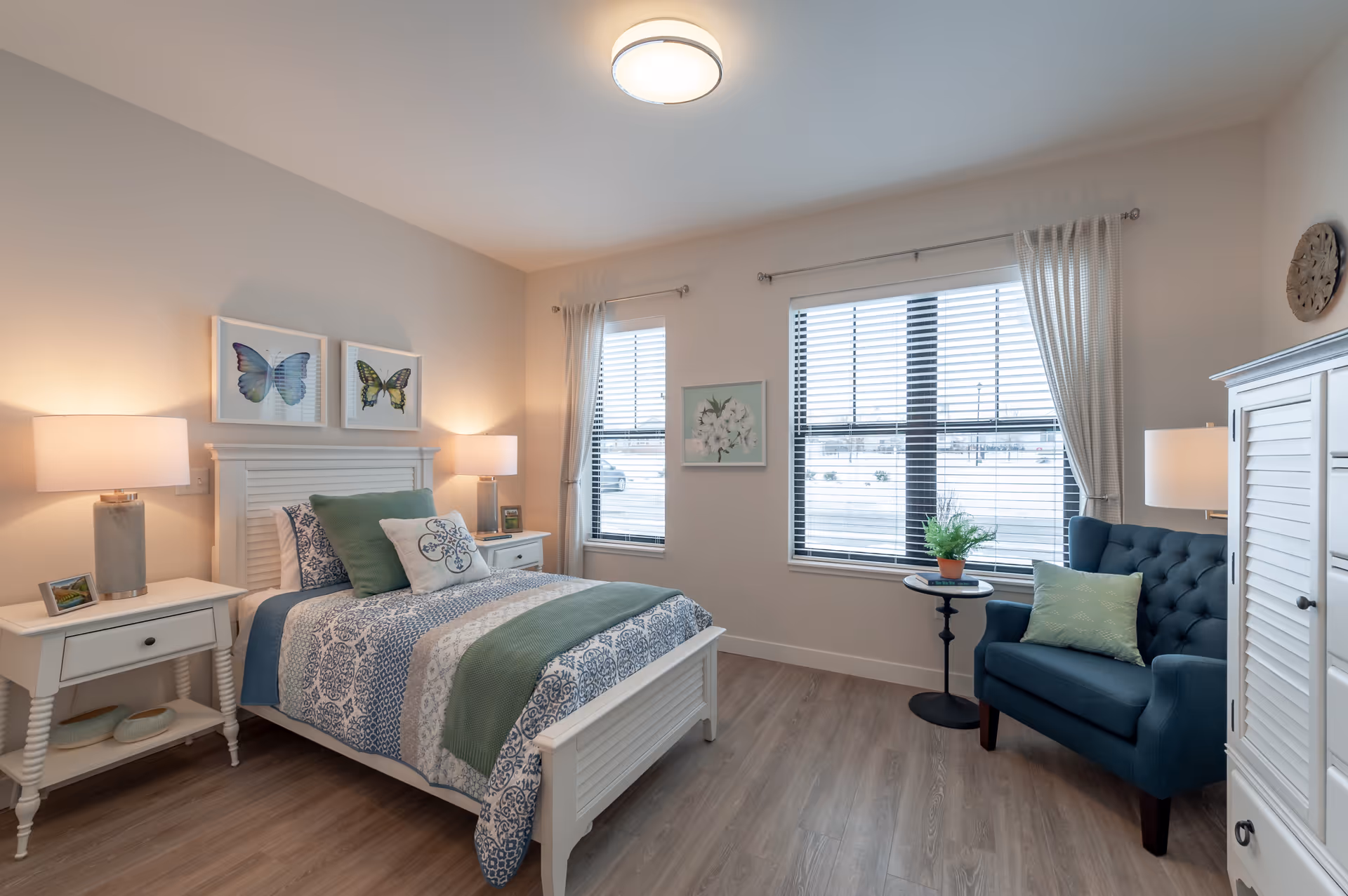 A bright and cozy bedroom with a white wooden bed adorned with blue and green bedding and pillows. Two bedside tables with lamps flank the bed. The room has two windows with white blinds and curtains, a blue armchair with a green pillow, a small round side table with a potted plant, and light wood flooring. Butterfly artwork hangs above the bed and a floral picture is on the wall between the windows.