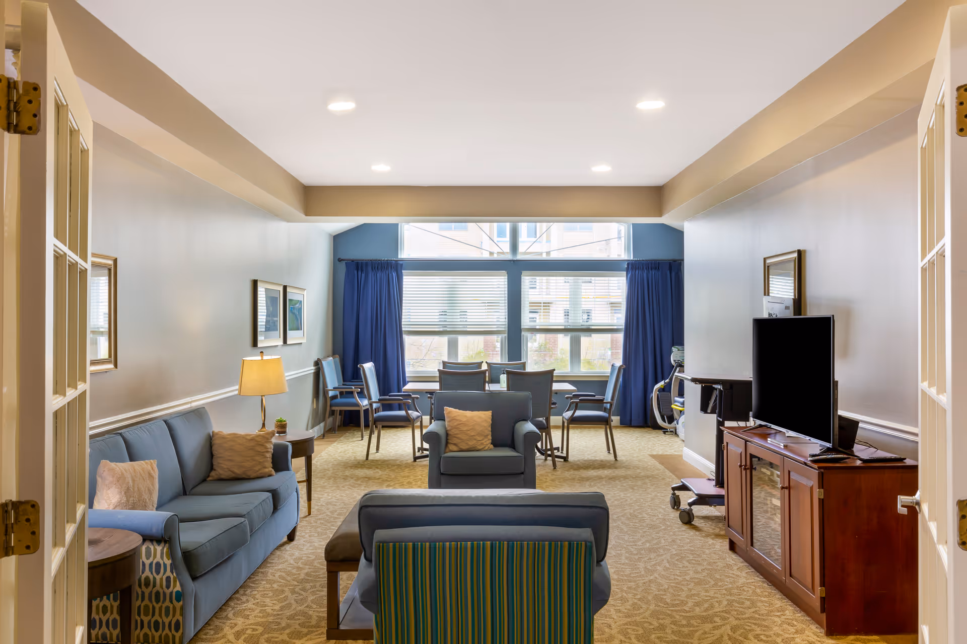 Bright communal living room with sofas, armchairs, a TV cabinet and a small dining area by large windows.
