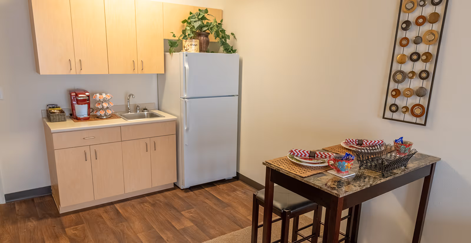 Small kitchen area with light wood cabinets, a white refrigerator, a sink, and a red coffee maker on the countertop. Next to the kitchen is a small dining table set for two with colorful plates, cups, and napkins. A decorative wall hanging with circular designs is on the wall above the table.