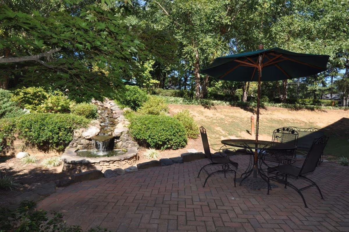 Outdoor patio area with a round metal table, four metal chairs, and a large green umbrella. The patio is paved with bricks and surrounded by greenery including bushes, trees, and a small stone waterfall feature.
