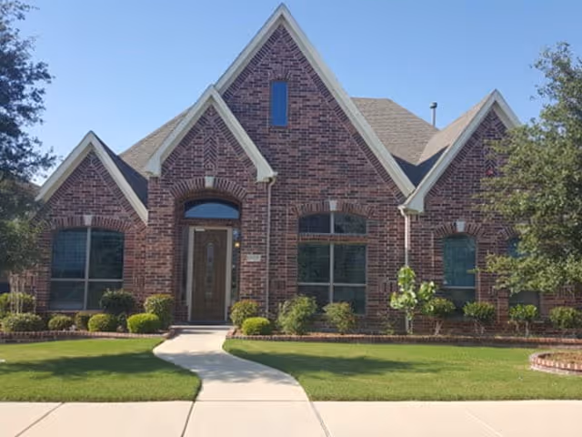 Front exterior view of a brick building with a steeply pitched roof, multiple gables, large windows, a central wooden door with glass panels, surrounded by neatly trimmed bushes and a green lawn under a clear blue sky.