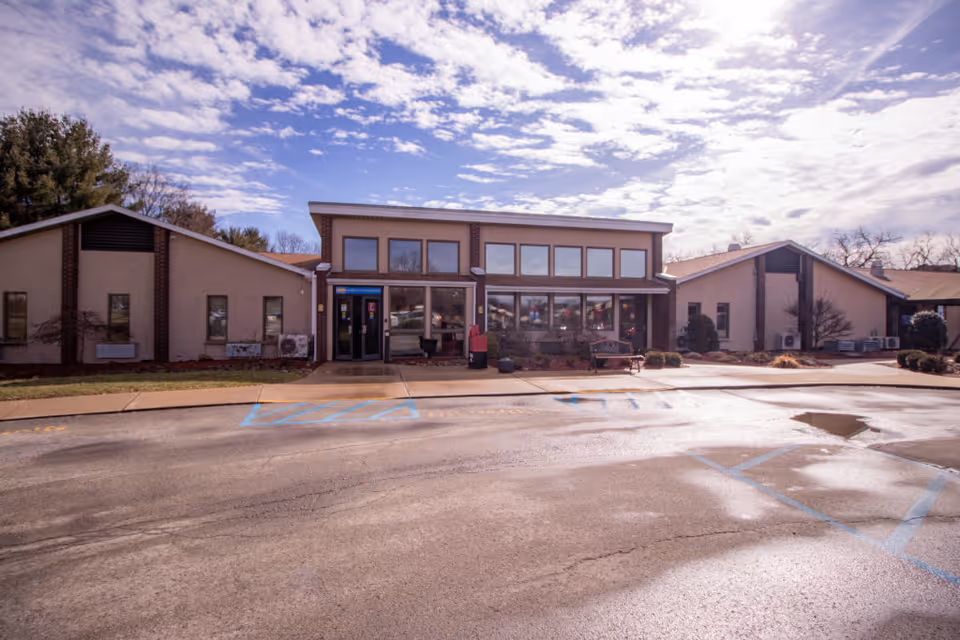 Exterior view of a single-story building with large windows and a central entrance, surrounded by a parking lot with marked handicapped parking spaces under a partly cloudy sky.