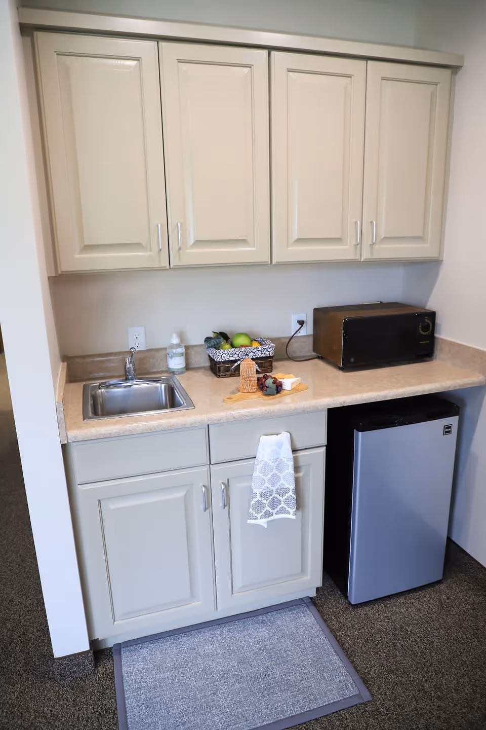 A small kitchenette area with beige cabinets, a stainless steel sink, a countertop with a basket of fruit, a cutting board with cheese and grapes, a microwave, and a mini refrigerator. A patterned towel hangs from one of the lower cabinet handles, and a gray floor mat is placed in front of the kitchenette.