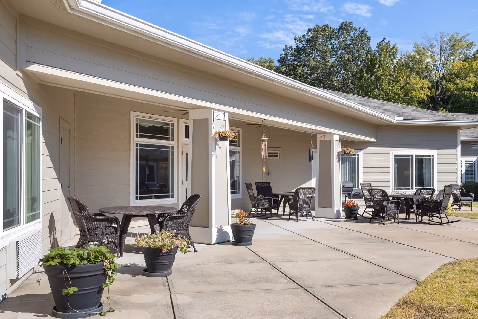 Outdoor patio area at Quail Ridge Assisted Living & Memory Care with multiple round tables and wicker chairs arranged for seating. Several potted plants and hanging planters decorate the space. The building exterior is light-colored with large windows, and trees are visible in the background under a clear blue sky.