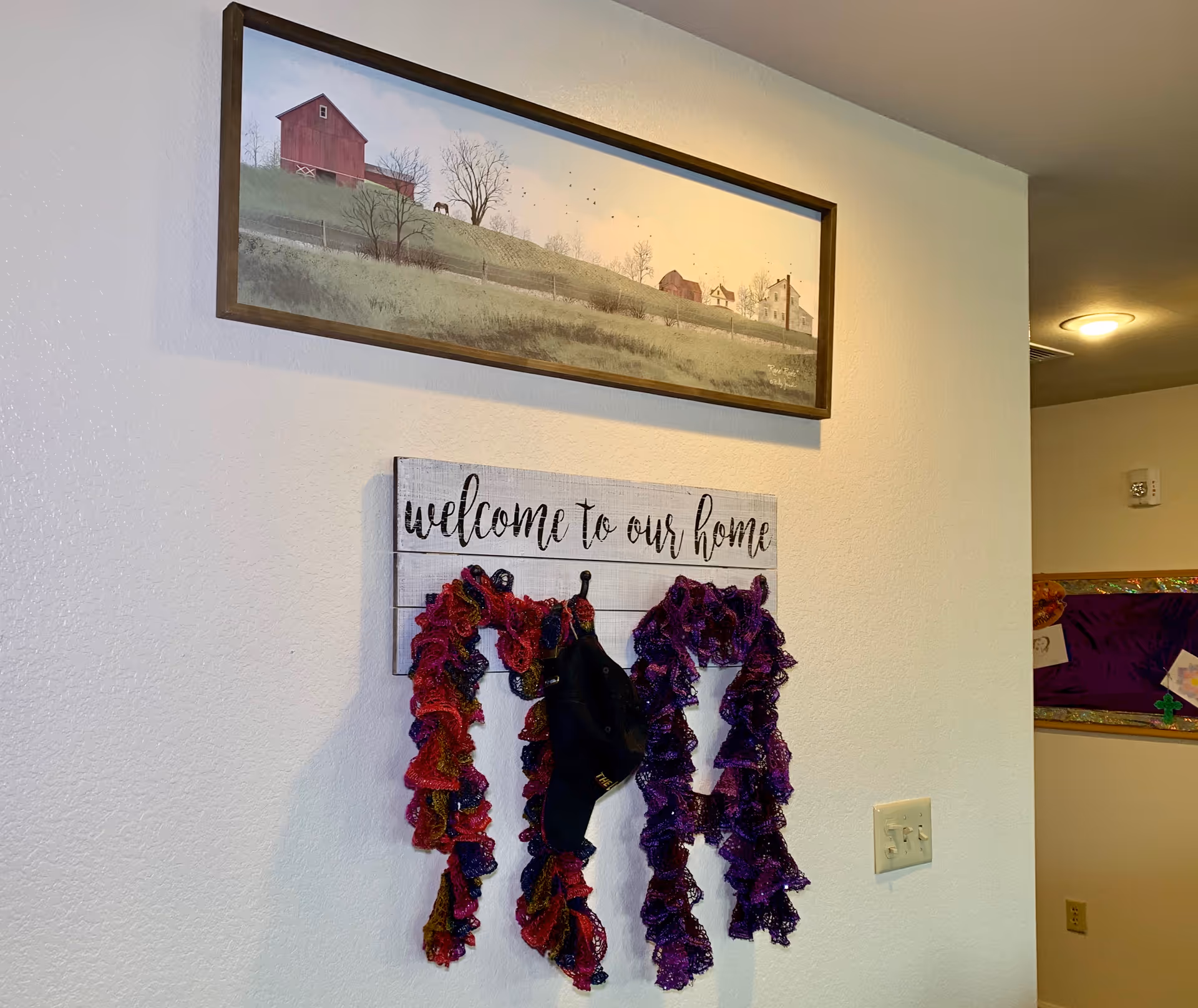 Interior wall with a framed landscape painting of a rural scene featuring a red barn and houses on a hill. Below the painting is a wooden sign that reads 'welcome to our home' with two colorful scarves and a black cap hanging on hooks attached to the sign. A light switch and part of a bulletin board with purple fabric and decorations are visible on the adjacent wall.