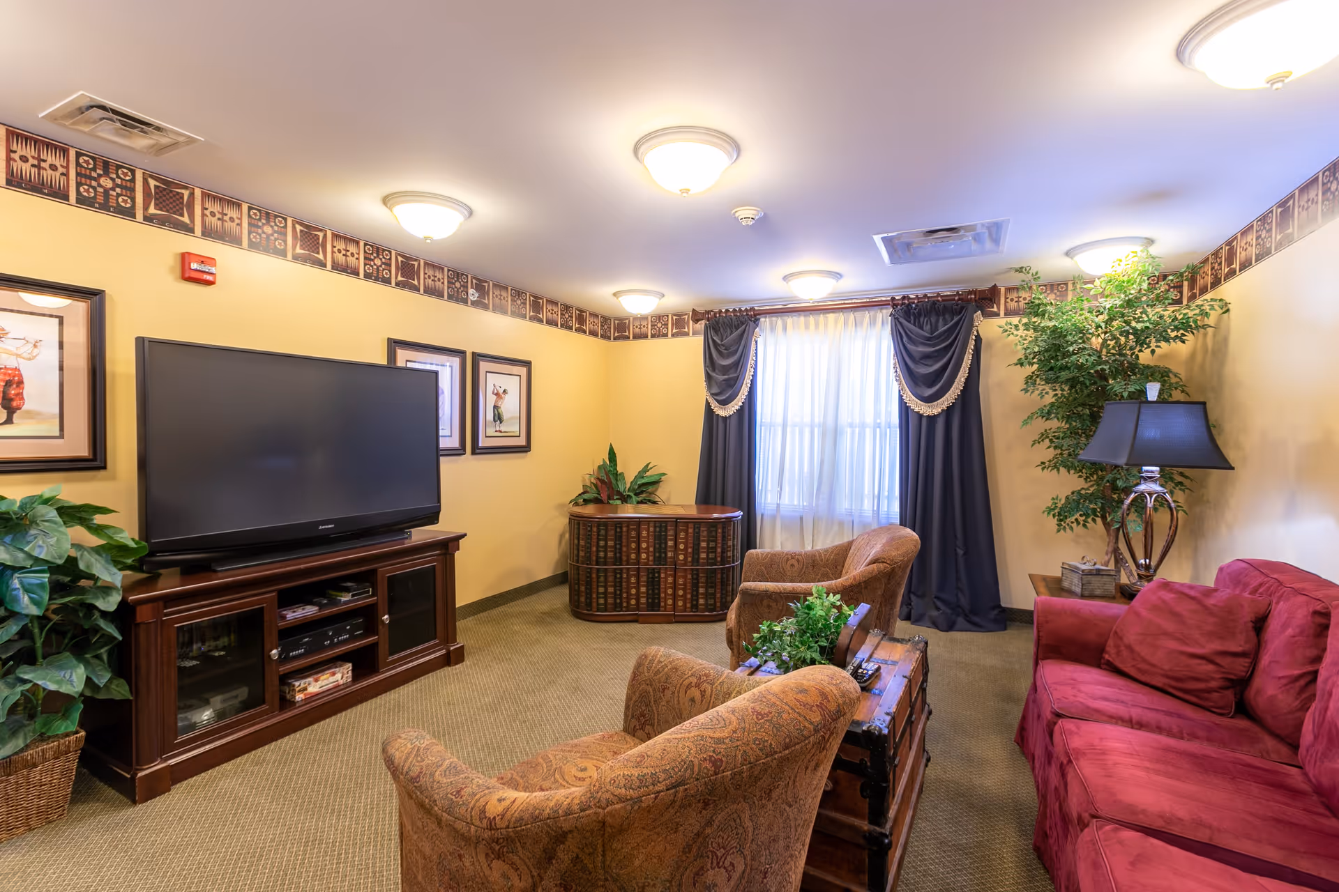 A cozy living room with a large flat-screen TV on a wooden stand, two patterned armchairs, a red sofa, a wooden chest used as a coffee table, a table lamp, framed artwork on the walls, and a window with dark curtains and sheer white drapes. The room has beige walls with decorative border trim near the ceiling and carpeted floor.