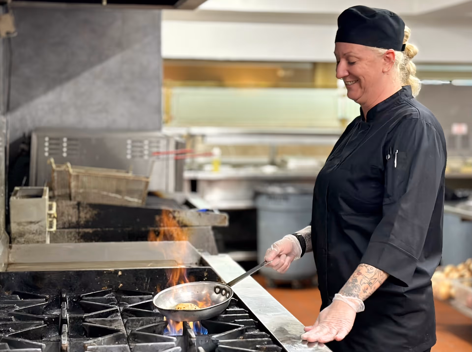 A chef wearing a black uniform and hat is cooking with a frying pan over a gas stove with visible flames in a commercial kitchen. The chef is smiling and wearing gloves, and the kitchen background includes industrial cooking equipment and utensils.