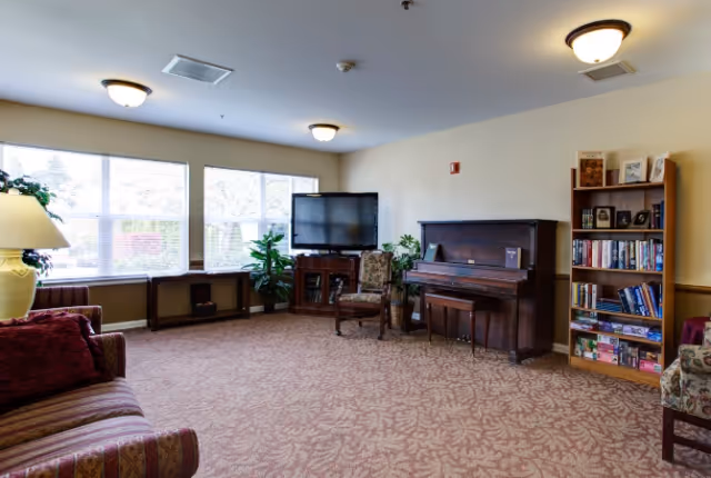 Communal living room with sofa, piano, television, bookshelf, chairs, and large windows.