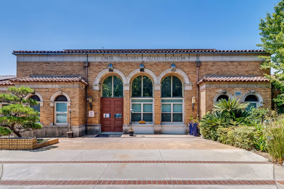 Front exterior view of a brick building with three large arched windows and a central red door. The building has a tiled roof and is surrounded by greenery including bushes and a small tree. Above the windows, the sign reads 'Emil Brown Auditorium'.