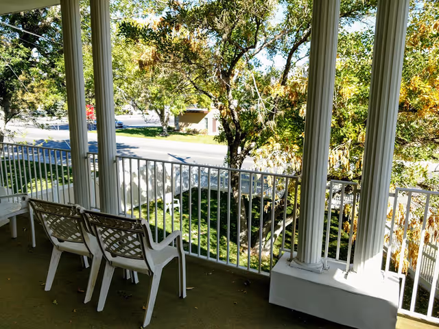 View from a covered balcony or porch with white railings and two white plastic chairs facing outward. The scene overlooks a green lawn with trees and a street in the background under a sunny sky.