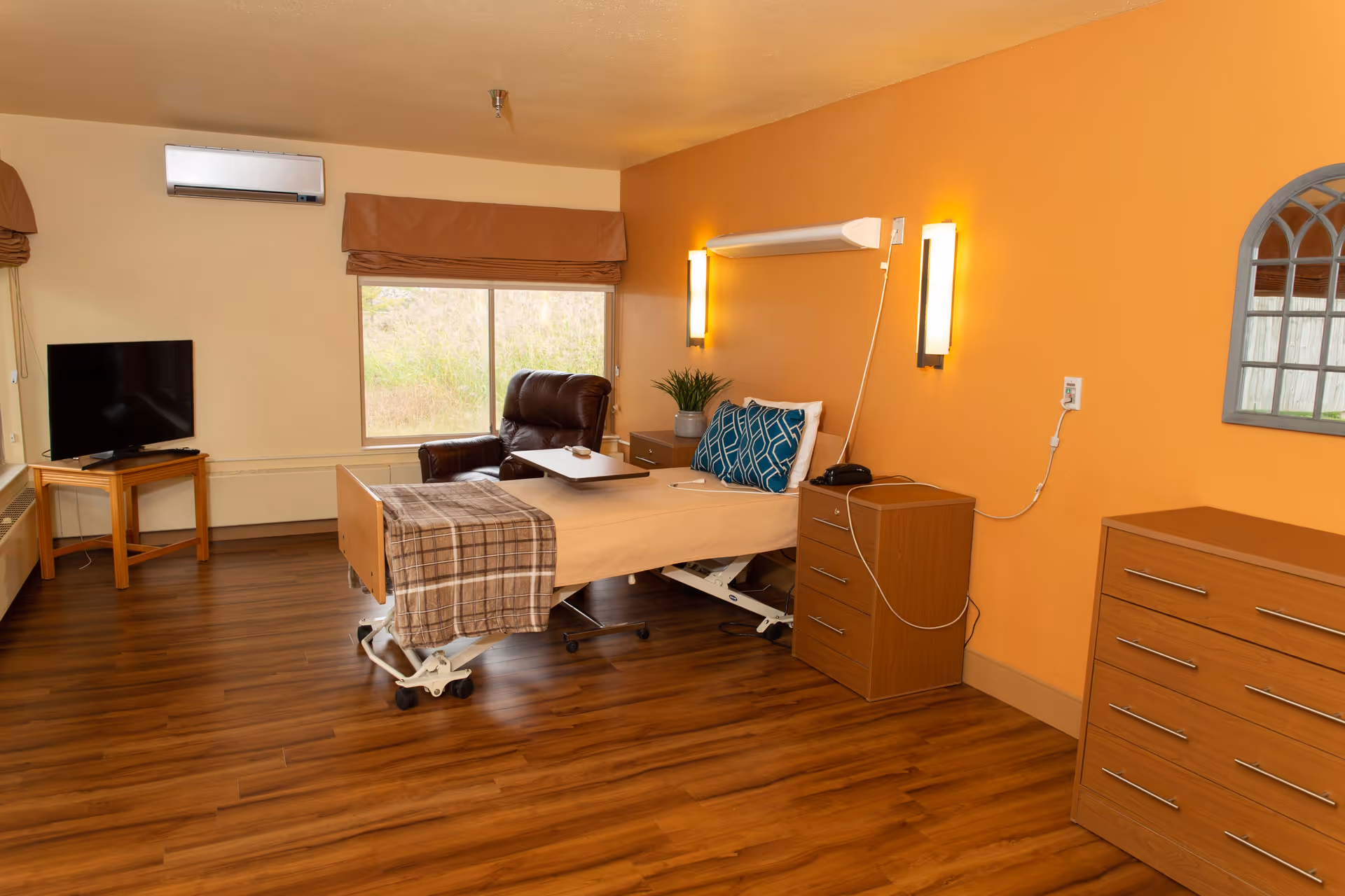 A senior living facility bedroom with a hospital-style bed covered with a beige blanket and a plaid throw. The bed is positioned near an orange accent wall with two wall-mounted lights and a small bedside table with a phone and a plant. There is a brown recliner chair next to a window with brown valances. A flat-screen TV sits on a wooden stand in the corner, and a wooden dresser is against the wall. The floor is wood laminate.