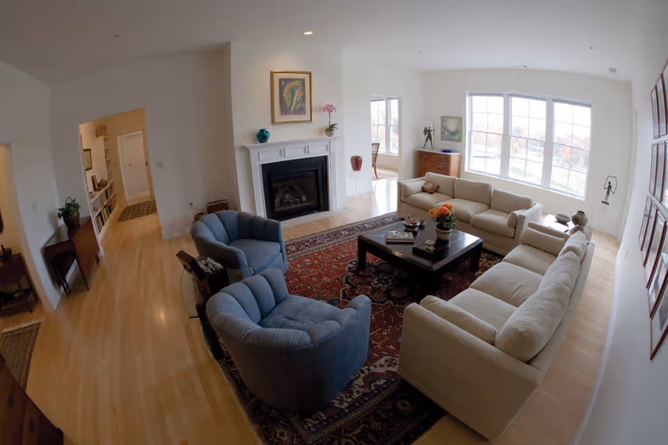 A bright living room with large windows letting in natural light. The room features two beige sofas and two blue armchairs arranged around a dark wooden coffee table on a patterned rug. There is a white fireplace with decorative items on the mantel, and artwork hanging above it. The room has light wood flooring and white walls, with additional furniture and decor visible in the background.