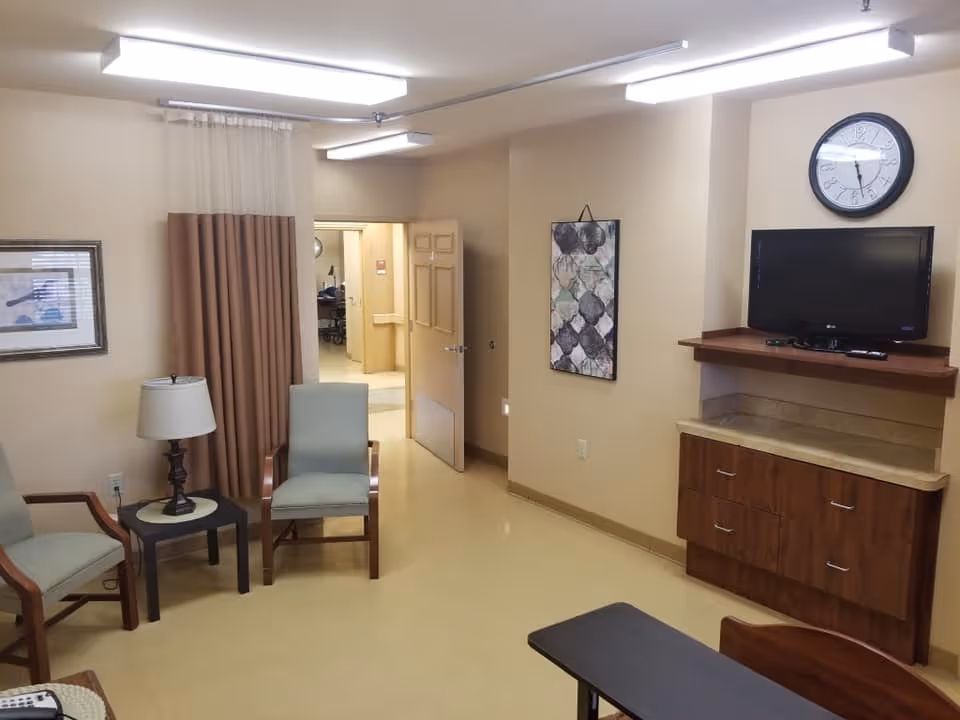 A small common area in a nursing facility with two armchairs, a side table with a lamp, a wall-mounted TV above a wooden cabinet, a wall clock, and abstract artwork on the wall. The room has beige walls and flooring, with an open door leading to a hallway.