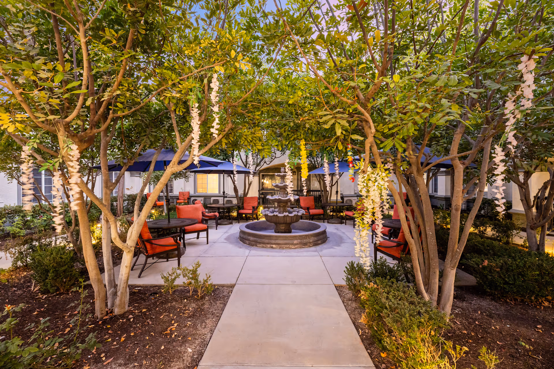 Outdoor courtyard area with a central multi-tiered water fountain surrounded by trees and hanging floral decorations. There are several round tables with red cushioned chairs and blue umbrellas arranged around the fountain. The area is paved with concrete walkways and bordered by landscaped bushes and plants.