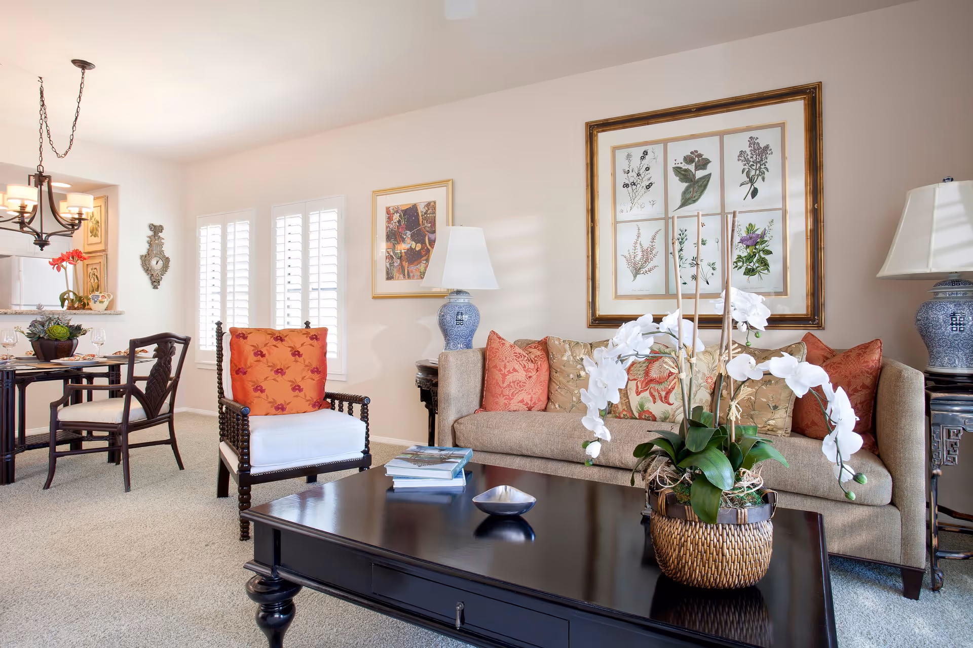 A bright and cozy living room area featuring a beige sofa with multiple patterned cushions, a dark wooden coffee table with a decorative plant and books, and a wooden armchair with a white cushion and an orange floral pillow. The room has light-colored walls adorned with framed artwork and a table lamp with a blue and white ceramic base. In the background, there is a dining area with a round table, chairs, and a chandelier hanging from the ceiling.